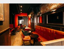 Schmaler Bar- oder Lounge-Innenraum mit roten Polsterbänken, kleinen runden Tischen und gemischten Sesseln auf Holzboden. Schwarze Wandverkleidungen mit Spiegeln, sichtbare Ziegelwand und dunkle Decke mit Spotbeleuchtung sowie ein Vorhang im hinteren Bereich prägen den Raum.
