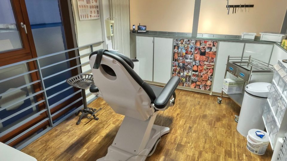 Behandlungsraum in einer Praxis mit höhenverstellbarem Stuhl in der Mitte, Holzfußboden und freiliegender Ziegeldecke mit Leuchtstofflampe. Entlang der Wände stehen Schränke und Arbeitsflächen mit Geräten sowie ein Fensterbereich mit Geländer.