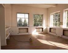 Heller, weitgehend leerer Innenraum mit Holzboden, weißen Wänden und mehreren großen Sprossenfenstern, durch die Bäume zu sehen sind. Unter den Fenstern sind eingelassene Heizkörper, und Sonnenlicht wirft lange Schatten in den Raum.