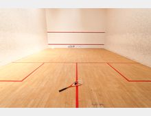 Ein Squashplatz mit Holzboden und roten Spielfeldmarkierungen, umgeben von weißen Wänden. Auf dem Boden liegt ein Squashschläger und ein schwarzer Squashball.