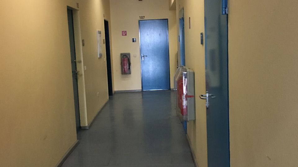Langer, schmaler Innenflur mit beige gestrichenen Wänden und grauem Boden, beleuchtet durch rechteckige Deckenleuchten. Entlang des Gangs befinden sich mehrere blaue Türen, am Ende eine Tür mit der Nummer „107“ sowie ein Feuerlöscher und ein Wandhydrant.