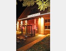 Außenansicht einer historischen Backsteinhalle mit der leuchtenden Beschriftung „Reithalle“ über dem Eingang. Der Eingangsbereich ist durch Glas und Metallstrukturen überdacht, und davor stehen einige Tische und Stühle auf einem gepflasterten Vorplatz, beleuchtet durch warme Lichter.