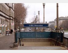 Eingang zur U-Bahn-Station "Weberwiese" in Berlin, mit einem Treppenabgang in die Station. Im Hintergrund befinden sich Gehwege, Gebäude, Bäume ohne Laub und die Fernsehturm-Spitze ist über den Bäumen sichtbar.