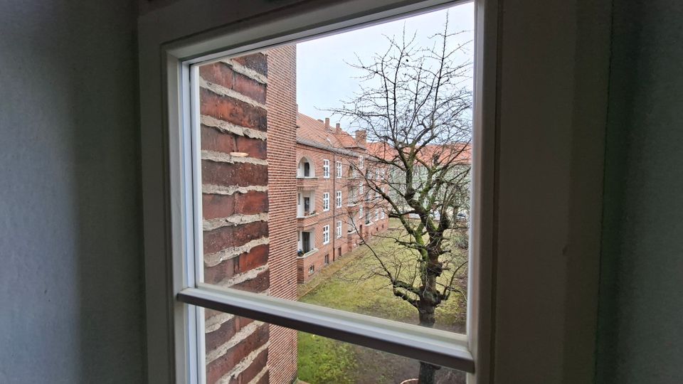 Blick durch ein weiß gerahmtes Fenster auf einen Innenhof mit Rasenfläche und einem kahlen Baum. Gegenüber stehen mehrstöckige Backstein-Wohnhäuser mit weißen Fenstern und roten Ziegeldächern.