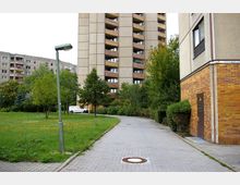 Wohnkomplex mit mehrstöckigen Plattenbauten, umgeben von einer Grünfläche mit Bäumen. Im Vordergrund führt ein gepflasterter Weg an einem Laternenpfahl vorbei, rechts ist ein Teil eines Gebäudes mit einer verklinkerten Fassade sichtbar.