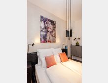 Modernes Schlafzimmer mit Doppelbett in weißer Bettwäsche und zwei korallfarbenen Kissen, flankiert von kleinen Nachttischlampen. Über dem dunklen Kopfteil hängt ein großes, farbiges Kunstbild; von der Decke hängen drei zylindrische Pendelleuchten, daneben steht ein Sideboard mit Vase.