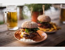 Zwei Teller mit Burgern aus Sesambrötchen, Salat, Käse, Speck und Beilage aus Pommes Frites stehen auf einem Holztisch, daneben befinden sich gefüllte Biergläser. Im Hintergrund ist unscharf eine Topfpflanze erkennbar.