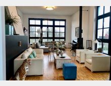 Wohnzimmer mit modernem, offenen Design, großen, schwarzen Sprossenfenstern, Holzboden und stilvollen Möbeln, darunter weiße Sofas, ein blauer Hocker und ein Couchtisch mit Dekoration. Ein Gaskamin und ein schwarzes Klavier befinden sich im Hintergrund, während eine Leopardenskulptur im Vordergrund steht.