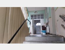 Treppenhaus in einem älteren Gebäude mit einem Geländer auf der linken Seite und einer mintgrünen Wand. Am oberen Ende der Treppe führt eine weiße Tür mit Glaseinsatz und Rundbogenfenster dahinter in einen weiteren Raum.