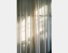 Halbtransparente, weiße Vorhänge vor einem Fenster, durch die warmes Sonnenlicht fällt. Die Fenstersprossen zeichnen sich als Schattenmuster auf dem Stoff ab.