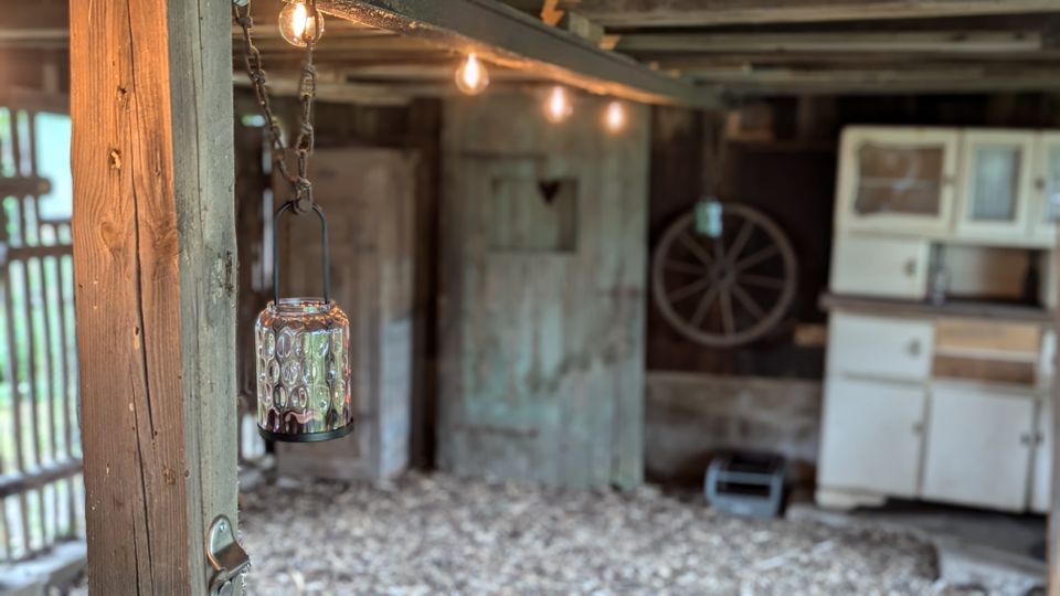 Innenansicht einer rustikalen Scheune mit groben Holzbalken, Kiesboden und einer hängenden Glaslaterne im Vordergrund. Im Hintergrund stehen alte Türen, ein Wagenrad an der Wand und ein weißer Schrank, darüber verläuft eine Lichterkette unter der Decke.