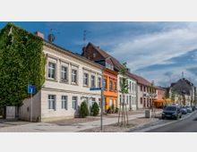 Straßenszene mit zweistöckigen Wohnhäusern in verschiedenen Farben, darunter weiße, orangefarbene und pastellgrüne Fassaden. Der Gehweg ist mit jungen Bäumen gesäumt, einige parkende Autos und ein Café mit Sonnenschirmen sind sichtbar.