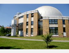 Backsteinbau mit großen Fenstern und halbrunder Kuppel aus Metall, vermutlich ein Planetarium, umgeben von einer Grünfläche. Im Vordergrund ein kleiner Fußweg und ein einzelner Baum, im Hintergrund blauer Himmel.