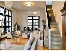 Offener Wohnraum mit moderner Einrichtung, großen schwarzen Fensterfronten und einer Treppe aus Beton an der rechten Wand. Der Raum ist mit Parkettboden, dekorativen Möbeln wie einem Sessel mit Zebramuster und einem niedrigen Couchtisch gestaltet.