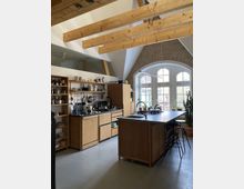 Offene Küche in einem Raum mit sichtbaren Holzdeckenbalken und einer Wand aus Backstein. Die Küche enthält eine Kochinsel mit Spüle, Holzschränke, offene Regale mit Geschirr sowie große Fenster mit abgerundeten Bögen, die viel Tageslicht hereinlassen.