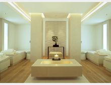 Minimalistischer Spa-Raum mit hellen Wänden, Holzboden und modernen Möbeln. Im Zentrum steht ein rechteckiger Steinbrunnen mit einer brennenden Kerze, dahinter ein Tisch mit einer Bonsai-Pflanze vor einer Wand mit strukturierter Verkleidung.