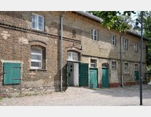Backsteinbau mit mehreren grünen Holzfensterläden und Türen, teils geöffnet. Über einer Tür ist ein Schild mit der Aufschrift „WC“ zu sehen. Der Vorplatz besteht aus Kopfsteinpflaster und der Bereich ist von Bäumen umgeben.