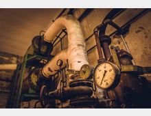 Detailaufnahme eines industriellen Rohrleitungssystems mit mehreren Rohren, Ventilen und einem Druckmesser. Die Umgebung wirkt alt und weist abgenutzte Wände und Metallstrukturen auf.