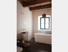 Ein minimalistisches Badezimmer mit einer freistehenden weißen Badewanne und einem passenden Waschbecken, dazu ein rustikaler Holzschemel. Die Wände sind schlicht gehalten, mit holzverkleideter Decke und einem Fenster mit schwarzem Rahmen, das Tageslicht hereinlässt.