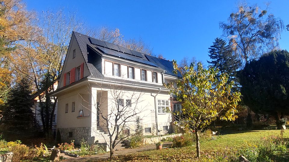 Zweistöckiges Wohnhaus mit hohem Satteldach und Solarpaneelen, umgeben von einem großen Garten mit Rasenfläche und Bäumen. Im Vordergrund sind ein kleiner Baum, Beete und ein schmaler Weg entlang der Hauswand zu sehen.