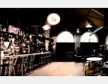 Innenraum einer Bar mit rustikalem Dekor. Auf der linken Seite befindet sich eine lange Theke mit Barhockern und Regalen voller Flaschen und Dekorationsobjekte. Auf der rechten Seite stehen Tische und Stühle, dahinter zwei große Fenster mit Rundbögen, die von Vorhängen teilweise verdeckt sind.