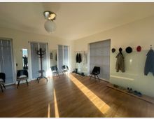 Ein heller Eingangs- oder Warteraum mit Holzboden, drei Fenstern mit Jalousien, mehreren schwarzen Stühlen, einem freistehenden Kleiderständer und einer Wand mit Haken für Kleidung und Hüte. Schuhe sind auf einer Holzablage am Boden platziert.