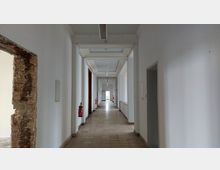 Langer, schlichter Flur in einem Gebäude mit weißen Wänden und hellbeigem Fliesenboden. An den Seiten befinden sich Zimmertüren, Feuerlöscher an der Wand und ein freiliegender Ziegelabschnitt links.