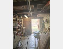 Innenansicht eines Geräteschuppens mit freiliegenden Holzbalken und Neonröhre an der Decke, Regalen mit Werkzeug und Gartenzubehör sowie einer Werkbank entlang der Wand. Eine große Holztür steht offen und gibt den Blick in einen Gartenbereich frei.