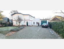 Einfamilienhaus mit weißer Fassade und grünen Türen, ergänzt durch rotbraune Ziegelakzente. Der gepflasterte Hof wird von einem geparkten Auto und einem kleinen Gartenbereich mit Hecken und niedrigen Pflanzen umgeben.