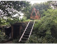 Eine bewaldete Umgebung mit einem kleinen, erdüberwachsenen Gebäude im Vordergrund, vermutlich ein Keller, daneben steht eine Metallleiter, die zu einer Holzschaukel mit Dreiecksgestell auf einer Anhöhe führt. Dichtes Laub umgibt die Szene.