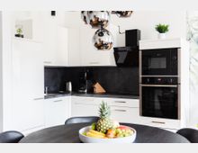 Moderne Küche mit weißen Hochglanzschränken, schwarzer Arbeitsplatte und eingebauten Elektrogeräten, darunter ein Backofen und eine Mikrowelle. Im Vordergrund steht ein dunkler Esstisch mit einer Obstschale, und eine auffällige Hängelampe mit verspiegelten Kugeln ist sichtbar.