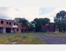 Verlassene Industriegebäude aus Ziegelstein mit zerbrochenen Fenstern und Graffiti, umgeben von überwuchertem Gelände und Bäumen. Der Asphaltboden ist teilweise mit Gras bewachsen, und der Himmel ist bewölkt.