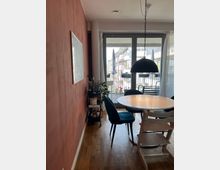 Esszimmer mit Holzboden und einer dunkelrot gestrichenen Wand links. Im Raum stehen ein runder Esstisch mit blauer Bestuhlung, darüber hängt eine schwarze Pendelleuchte. Im Hintergrund führen Glastüren auf einen Balkon mit Blick auf moderne Wohnhäuser.