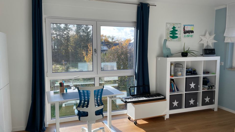 Helles Kinderzimmer mit einem großen Fenster, blauen Vorhängen und Blick auf eine grüne Landschaft. Im Raum stehen ein weißer Schreibtisch mit einem blauen Bürostuhl, ein Regal mit Spielzeug und Aufbewahrungsboxen sowie eine elektrische Tastatur auf einem Rollwagen.