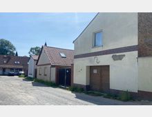 Eine ländliche Hofanlage mit mehreren Gebäuden, darunter ein verputztes Haus mit braunen Holztoren und bräunlichen Ziegelakzenten sowie kleinere Häuser mit roten Ziegeldächern. Im Hintergrund sind parkende Autos und Bäume sichtbar, während der Boden aus einer unbefestigten Kiesfläche besteht.