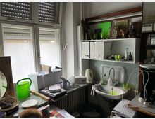 Kleiner, vollgestellter Arbeitsraum mit großen Fenstern und halb geschlossenen Rollläden, einer Fensterbank mit Ordnern, Gießkanne und Papieren. Rechts befindet sich ein Wandregal mit weiteren Ordnern und Gegenständen sowie ein kleines Waschbecken mit Armatur und abgestelltem Geschirr.