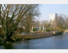 Flussufer mit gemauerter Uferbefestigung und einem Außenbereich mit Sonnenschirmen und Tischen, umgeben von Bäumen. Im Hintergrund ein modernes Hochhaus und weitere Gebäude, umgeben von einer parkähnlichen Landschaft.