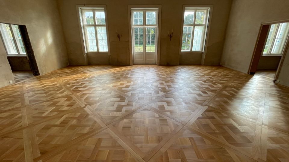 Großer, leerer Innenraum mit hellem, geometrisch gemustertem Parkettboden und hohen Wänden mit teilweise abblätternder Decke. An der Stirnseite befinden sich drei hohe Fenster und eine doppelflügelige Glastür zum Außenbereich, seitlich weitere Tür- und Fensteröffnungen.