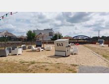 Sandfläche mit Strandkörben, weißen und grauen Kunststoffstühlen sowie Sonnenschirmen aus Stroh. Im Hintergrund befindet sich eine Steinmauer, moderne Gebäude und eine blaue Fußgängerbrücke unter bewölktem Himmel.