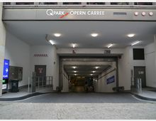 Einfahrt zu einem öffentlichen Parkhaus mit der Beschilderung "Q-Park Opern Carree". Der Eingang ist mit beleuchteten Deckenlampen, Hinweisschildern zu Höhen- und Zugangsbeschränkungen sowie einer Rampe ausgestattet.