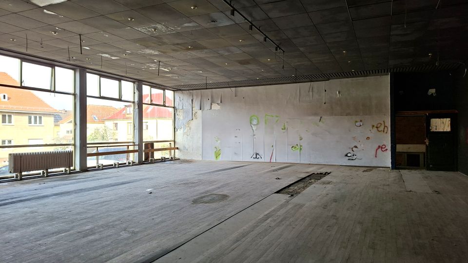 Leerer, verlassener Innenraum eines ehemaligen Gewerberaums mit großflächigen Fensterfronten zur Straße, abgenutztem Holzboden und dunkler Rasterdecke mit freihängenden Leitungen. An der Rückwand sind beschädigte Verkleidungen und Graffiti zu sehen, daneben ein offener Durchgang zu einem Nebenraum.