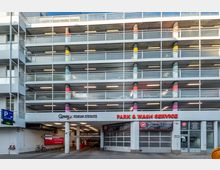 Mehrstöckiges Parkhaus mit einer Fassade aus Beton und Metallgeländern, gekennzeichnet durch bunt bemalte Säulen. Im Erdgeschoss befinden sich Eingänge zu einem Park- und Waschservice sowie Schilder mit Informationen zur Autowäsche und Parkmöglichkeiten.