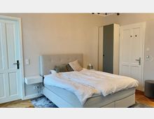 Ein Schlafzimmer mit einem grauen Polsterbett, weißer Bettwäsche und mehreren Kissen. Links befindet sich eine weiße Tür, daneben ein wandmontierter Nachttisch; rechts stehen ein grauer Schrank und eine weitere Tür. Der Raum hat einen Parkettboden.