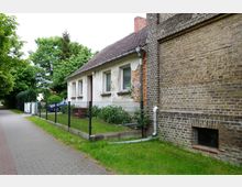 Ein einfaches Wohngebäude mit Putzfassade und Satteldach, angrenzend an eine gemauerte Backsteinwand. Im Vordergrund ein kleiner umzäunter Garten, daneben ein gepflasterter Gehweg und Bäume im Hintergrund.