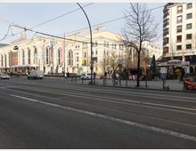 Straßenansicht mit dem Friedrichstadt-Palast, einem großen Gebäude mit schmuckvoller Fassade und Werbebannern, im Hintergrund. Im Vordergrund verlaufen Straßenbahnschienen und eine Kreuzung, umgeben von Gehwegen mit geparkten Fahrrädern, einem Imbissstand und Geschäften.