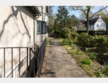 Ein gepflasterter Gehweg führt entlang einer Hauswand mit vergitterten Fenstern, umgeben von grünen Sträuchern und Blumenbeeten. Im Hintergrund sind weitere Wohnhäuser und große Bäume zu sehen.