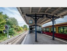 Überdachter Bahnsteig mit einem gelb-roten S-Bahn-Zug rechts und einer freien Gleisstrecke links, umgeben von Bäumen. Ein Schild mit der Aufschrift „Kurzzug Ende“ hängt an einer dunkelblauen gusseisernen Säule.
