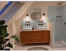 Modernes Badezimmer in einem Dachgeschoss mit Holz-Waschtisch, zwei rechteckigen Waschbecken und blauen Wandfliesen. Eine Glasdusche mit Regenduschkopf befindet sich rechts, und links steht eine große, grüne Pflanze auf einem schwarzen Gestell.