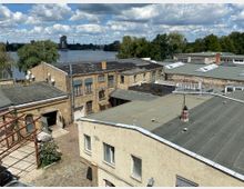 Mehrere flache Industriebauten mit Backstein- und Putzfassaden in einem Hof, umgeben von einem gepflasterten Boden. Im Hintergrund ist ein Fluss mit Bäumen und vereinzelten Hochhäusern unter einem bewölkten Himmel zu sehen.