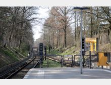 Bahnhof einer U-Bahn-Station in einer waldreichen Umgebung. Im Vordergrund ist der Bahnsteig mit Signalanlagen und Spiegeln zu sehen, im Hintergrund führen die Gleise in eine von Bäumen gesäumte Strecke.